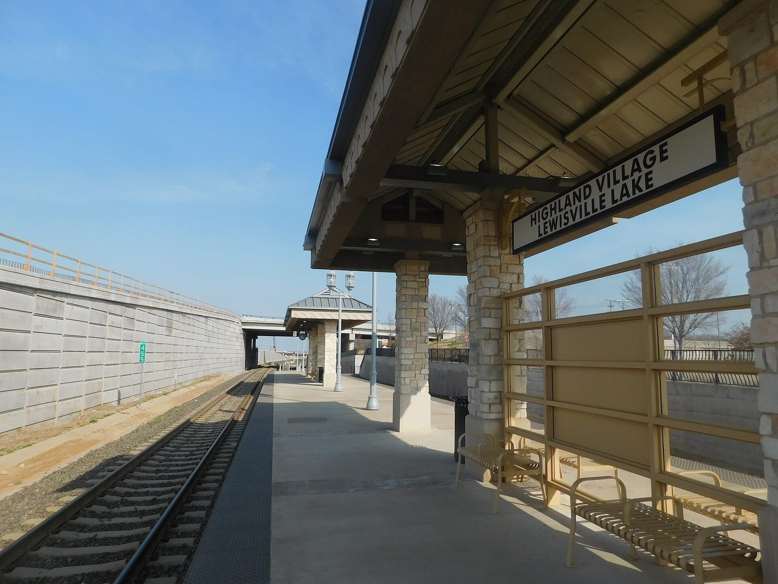 The Highland Village/Lewisville Lake A-train station platform, with limestone columns under a wood-ceiling canopy, the station name sign, and the track beside a concrete retaining wall