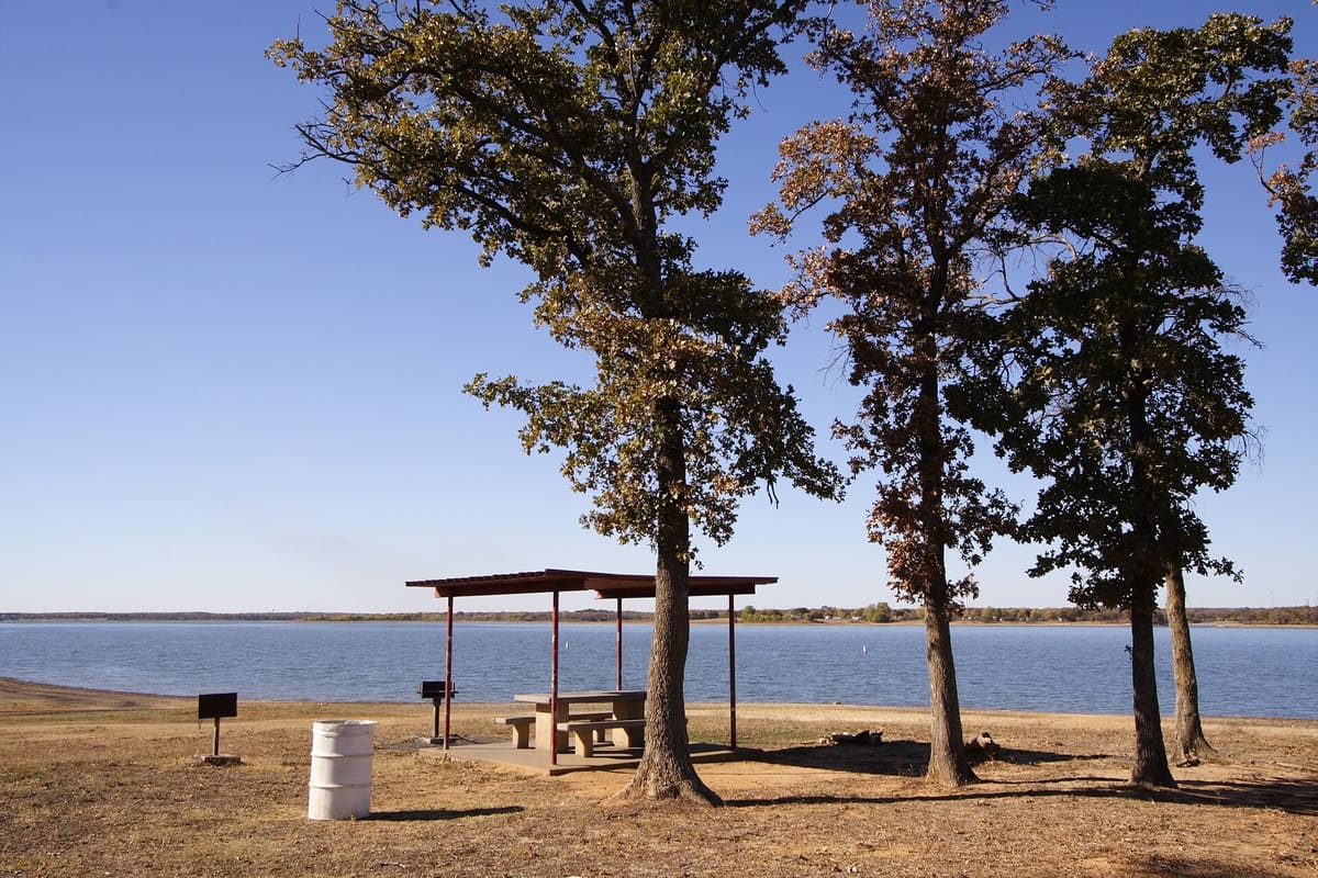 A covered picnic table beneath tall oak trees on a grassy Lewisville Lake shoreline, with blue water and a low wooded far shore
