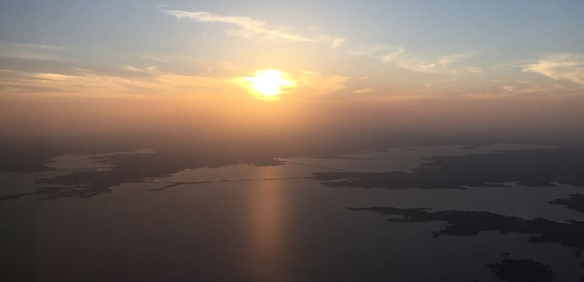 Lewisville Lake from the air at sunset, with the sun low over a hazy horizon, wooded shorelines and islands, and a long bridge crossing the water