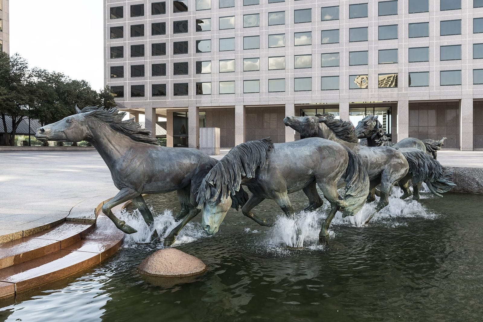 The bronze Mustangs of Las Colinas splashing through the fountain stream at Williams Square, with a granite office tower behind them