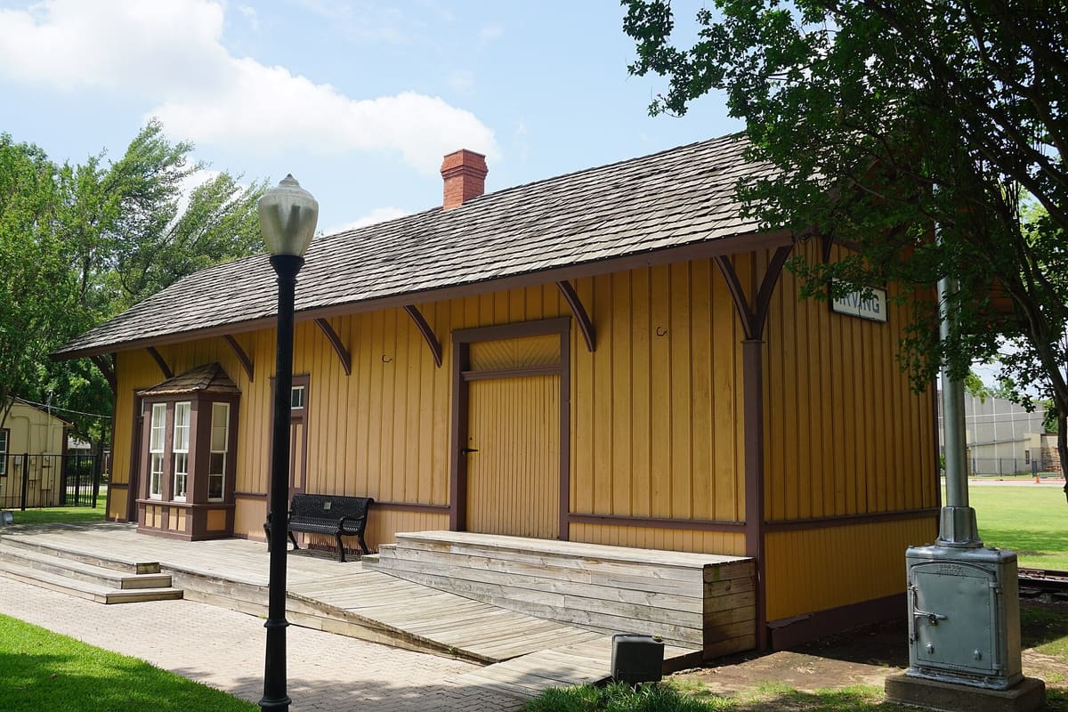The board-and-batten Heritage Park depot in downtown Irving, painted mustard yellow with brown trim, a bay window, and an Irving station sign