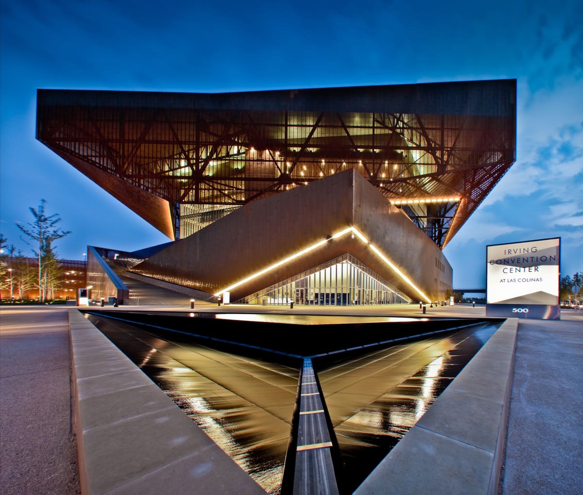 The Irving Convention Center at Las Colinas at dusk, its copper-clad upper volume cantilevered over a lit glass entrance and a reflecting pool