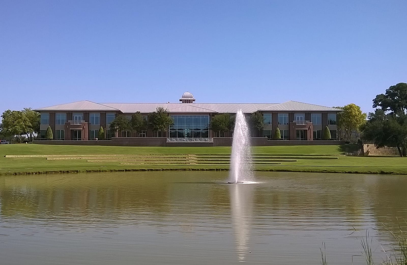 Keller Town Hall, a two-story brick building with a standing-seam metal roof and a domed cupola, seen across a lake with a fountain at The Parks at Town Center