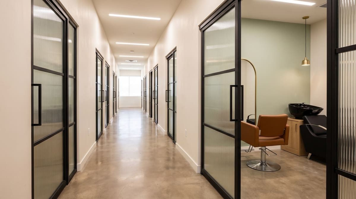 Finished salon-suites corridor with polished concrete floors, black-framed studio doors, and an open studio showing a cognac styling chair, arched brass mirror, and black shampoo bowl