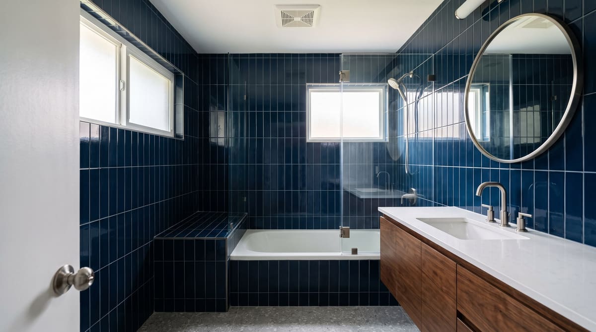 A remodeled 1970s hall bathroom with the original tub behind a clear glass panel, deep navy vertical stack tile, a gray terrazzo-look floor, and a floating walnut vanity