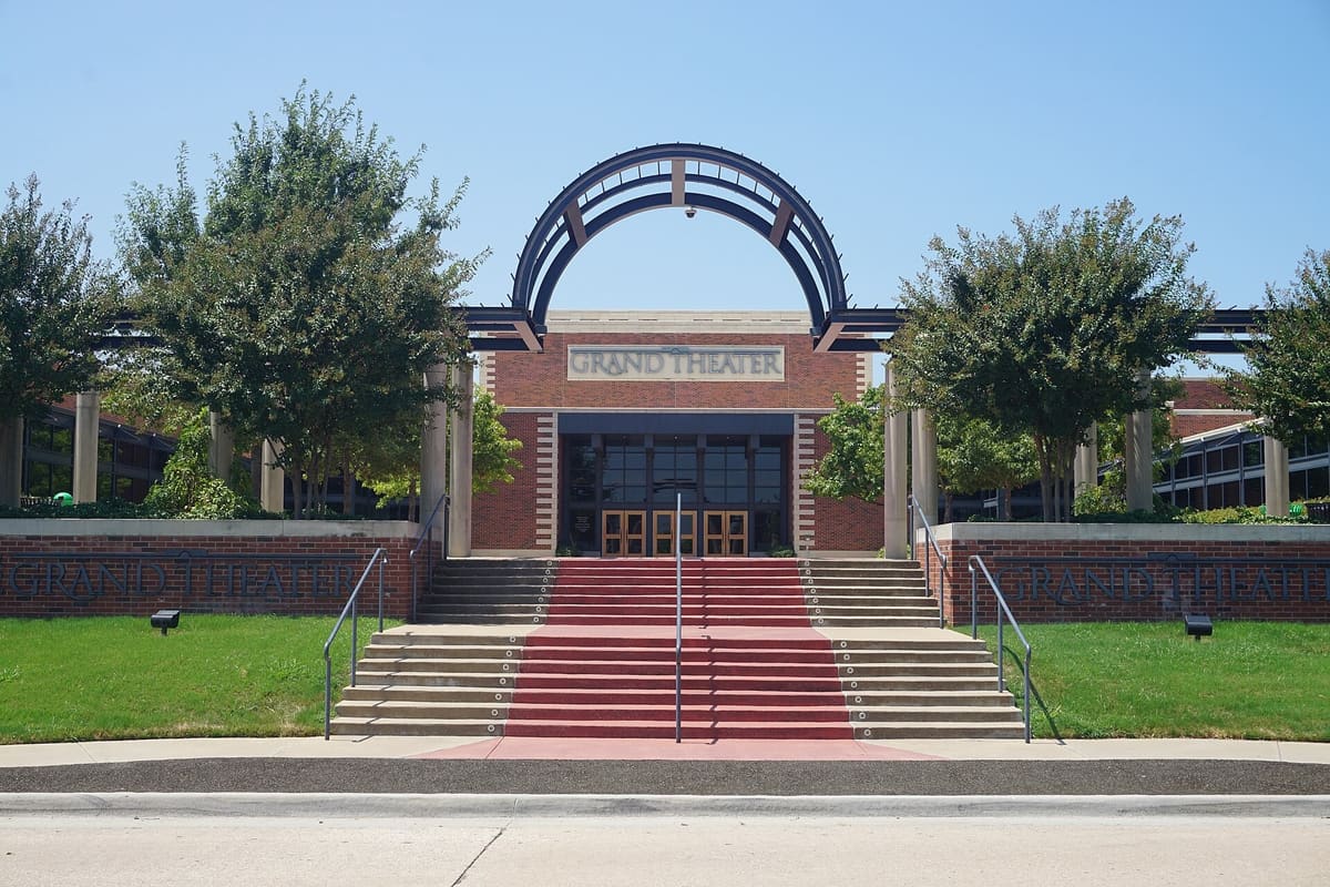The Grand Theater in Old Town Lewisville, a red brick entrance with a steel arch overhead, a red carpeted staircase, and trees on either side