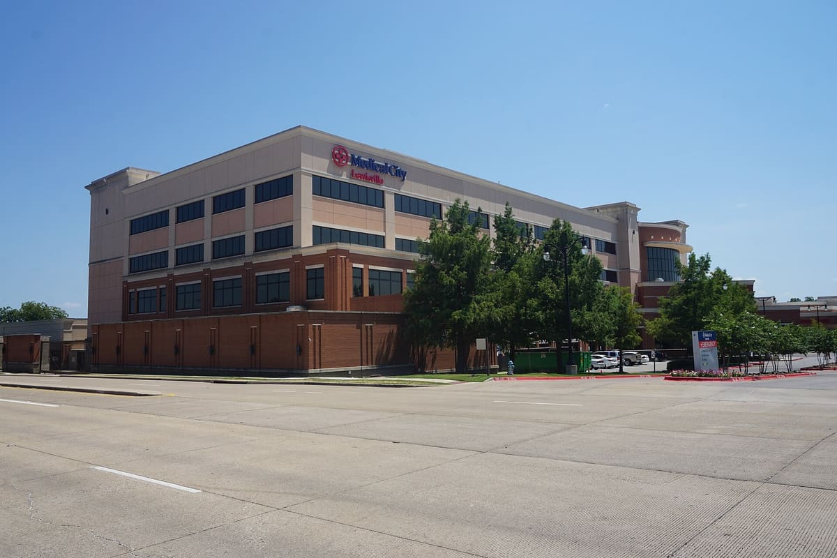Medical City Lewisville hospital, a multistory brick and stucco building with rows of windows and a red Medical City sign, seen across a wide street