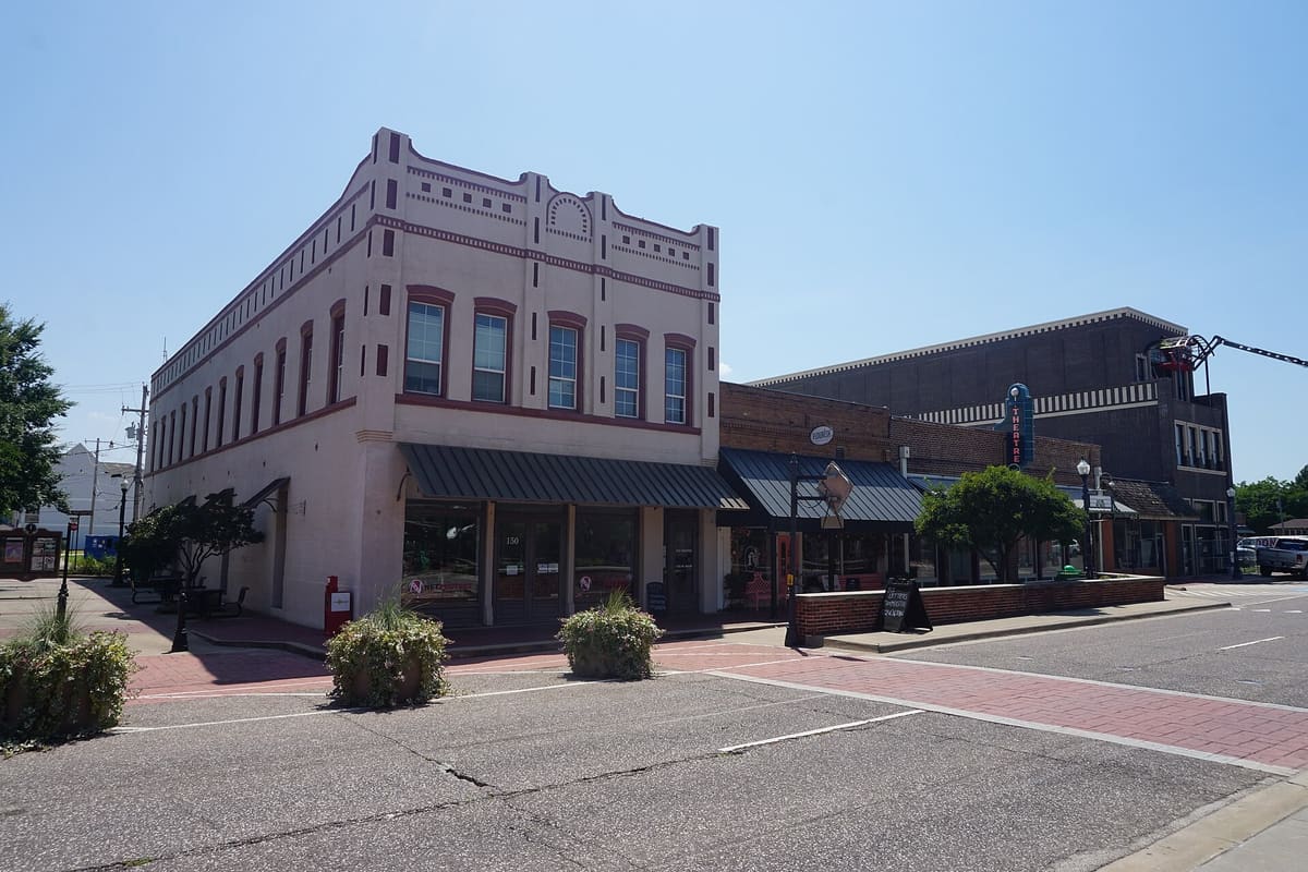 Old Town storefronts on West Main Street in Lewisville, a two-story white masonry commercial building with arched window hoods and a metal awning beside a row of brick storefronts