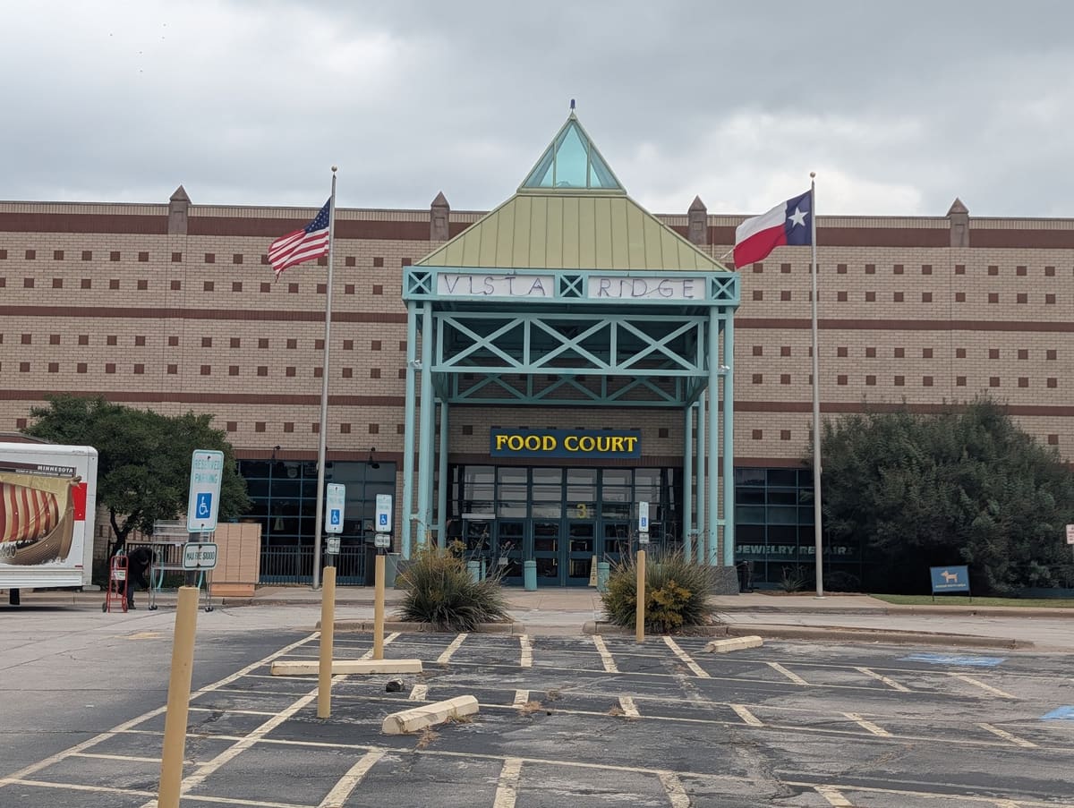 The Vista Ridge Mall food court entrance in Lewisville, a green steel and glass pavilion with a pyramid roof set into a long brick facade, with American and Texas flags out front