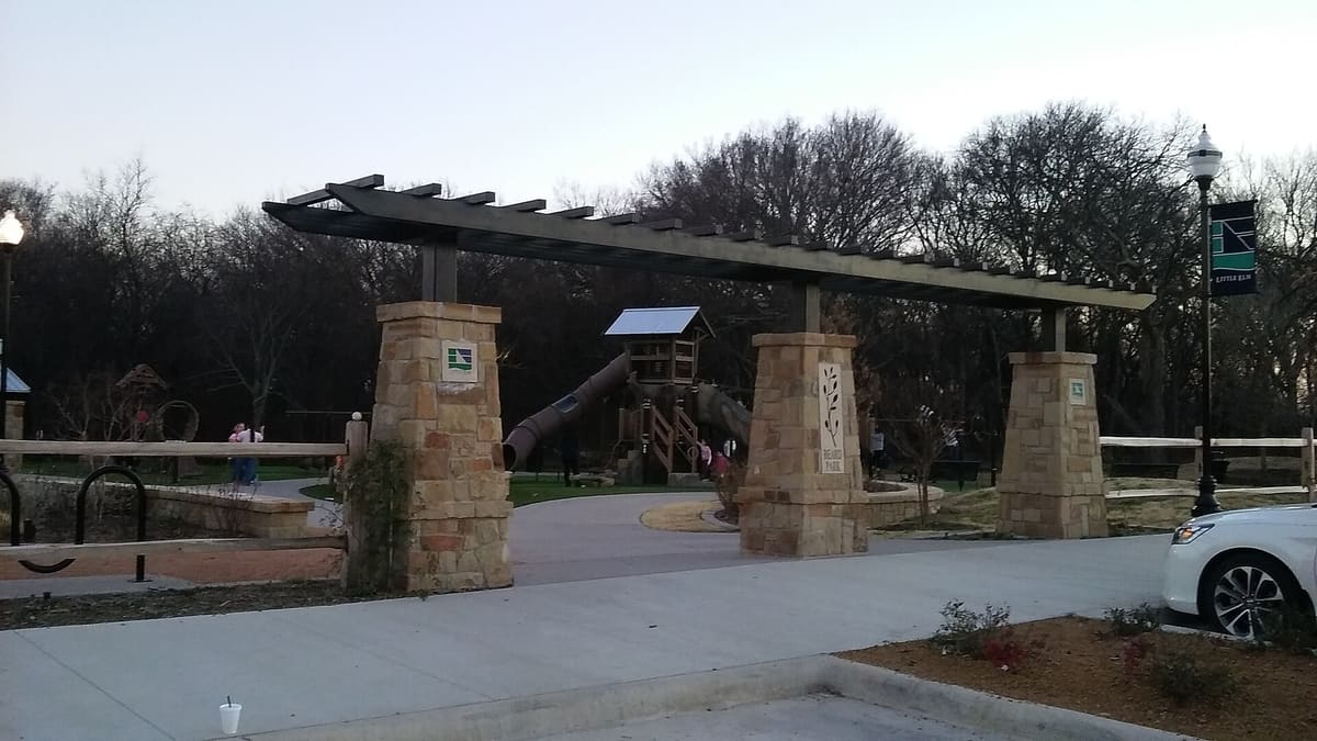 The stone-pillared entrance to Beard Park in Little Elm with a timber pergola, a Beard Park sign, and a wooden play tower behind it at dusk