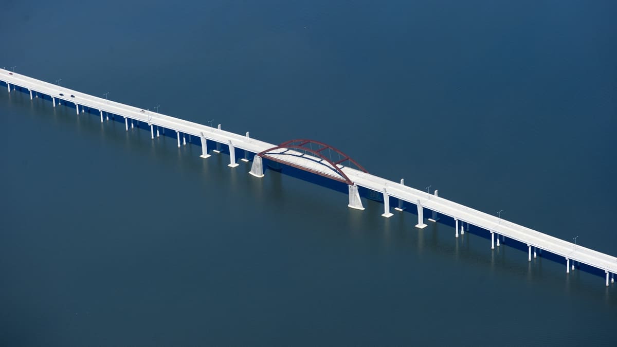 Aerial view of the Lewisville Lake Toll Bridge, a long white concrete causeway crossing dark blue water with a red steel arch at its center