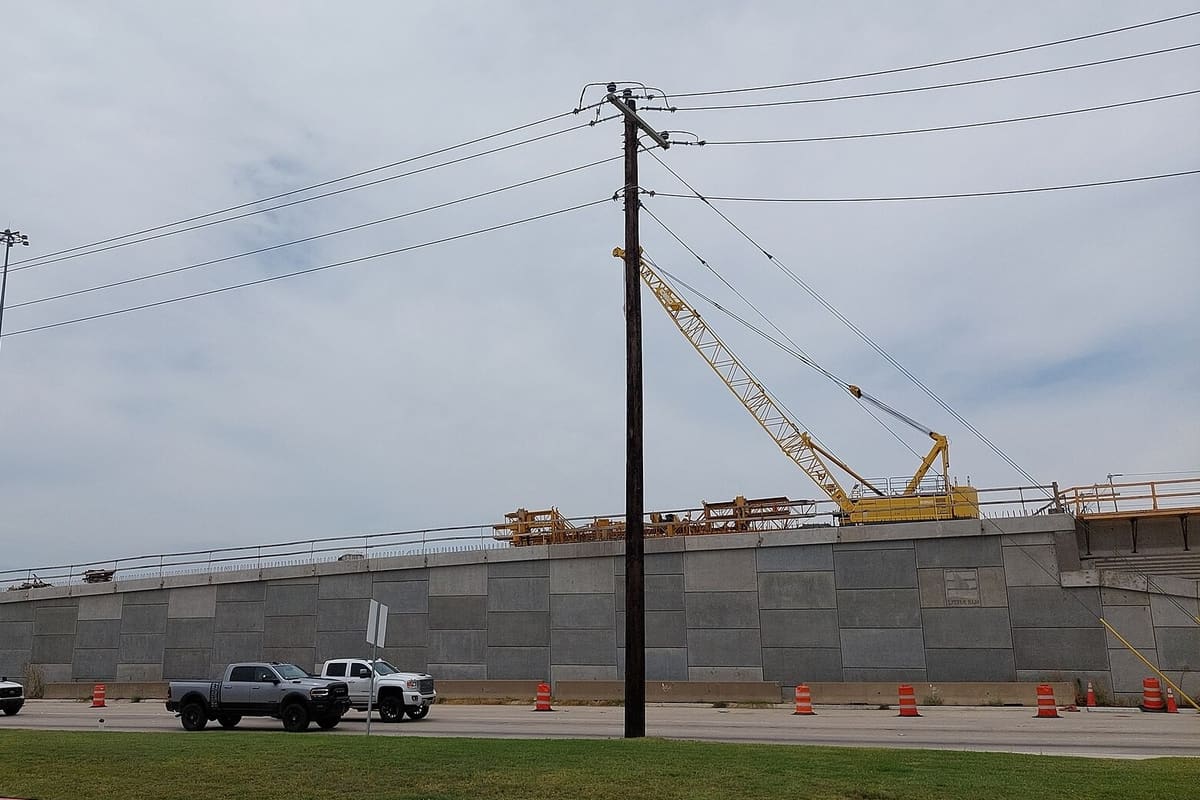 Highway overpass construction on US 380 at FM 720 in Little Elm, with a tall concrete retaining wall, a yellow crawler crane on the deck, and orange traffic barrels below