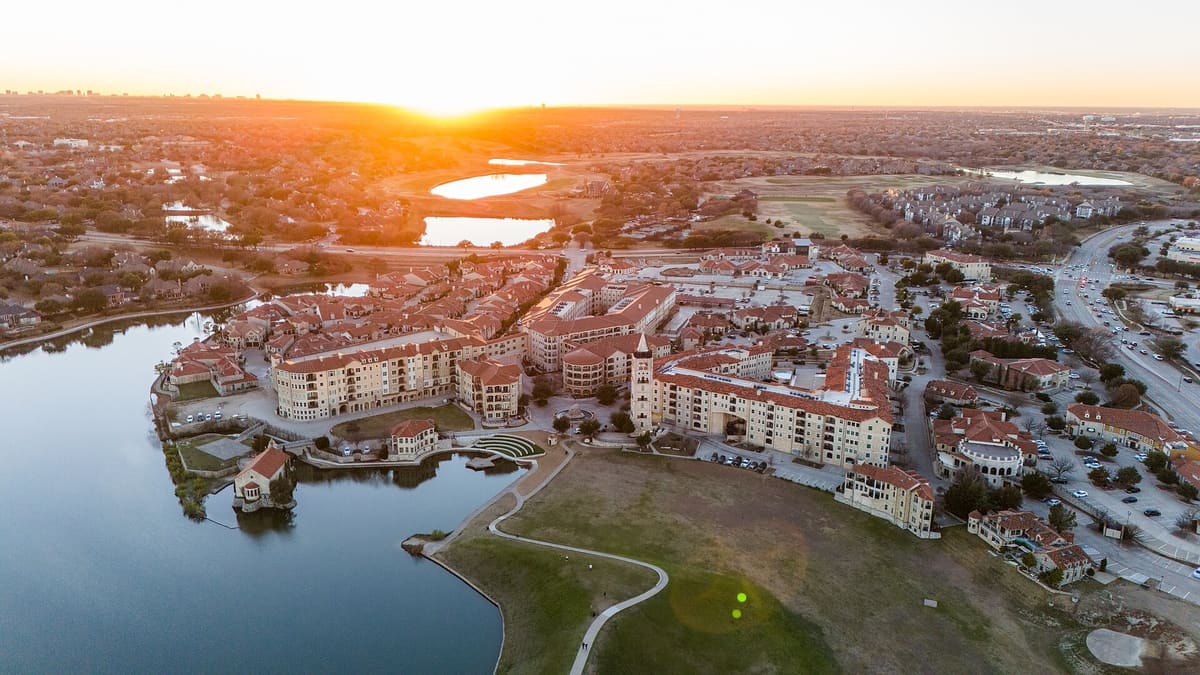 Aerial view of Adriatica Village in McKinney at sunset: stone buildings with red tile roofs, a bell tower, and a chapel on a small island in the lake