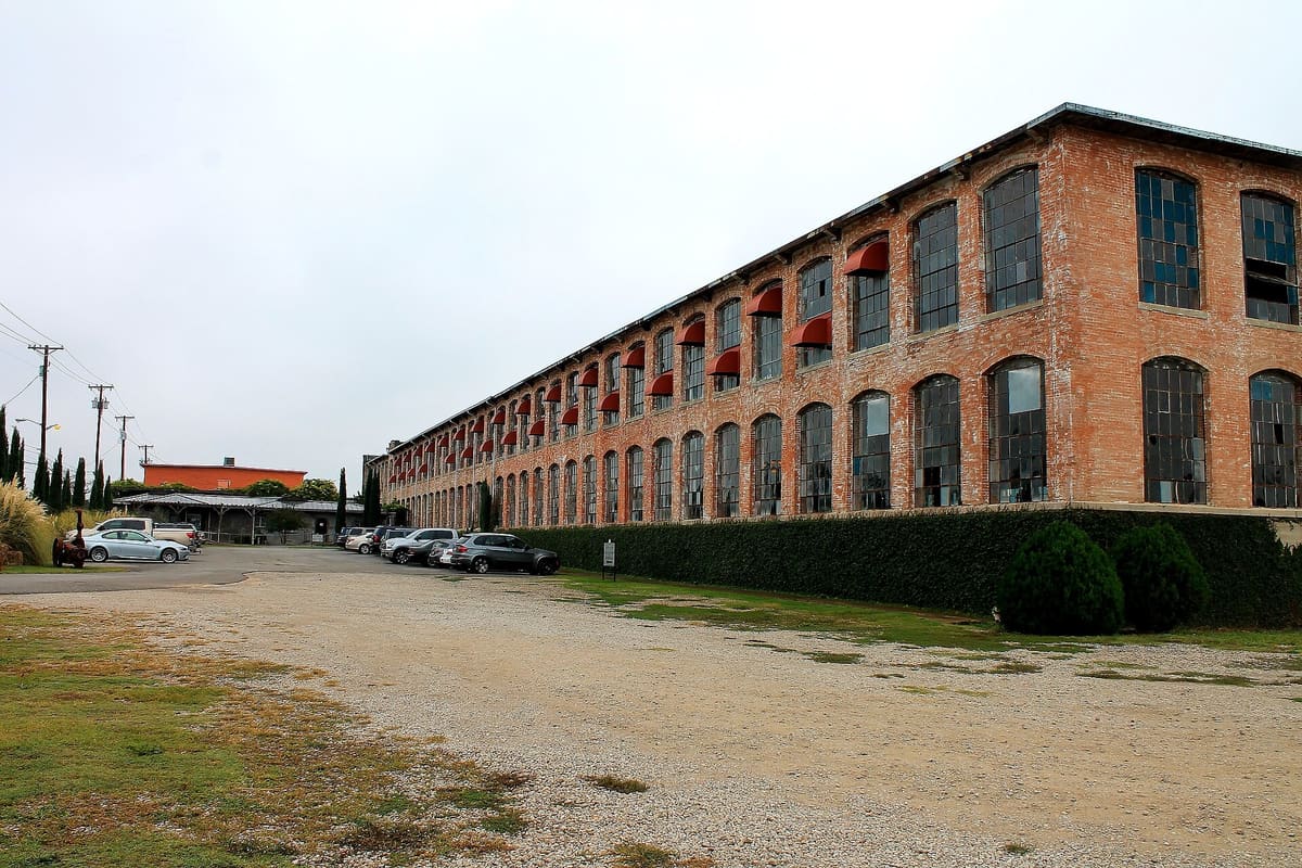 The McKinney Cotton Mill, a long two-story red brick mill building with rows of arched steel windows and red awnings