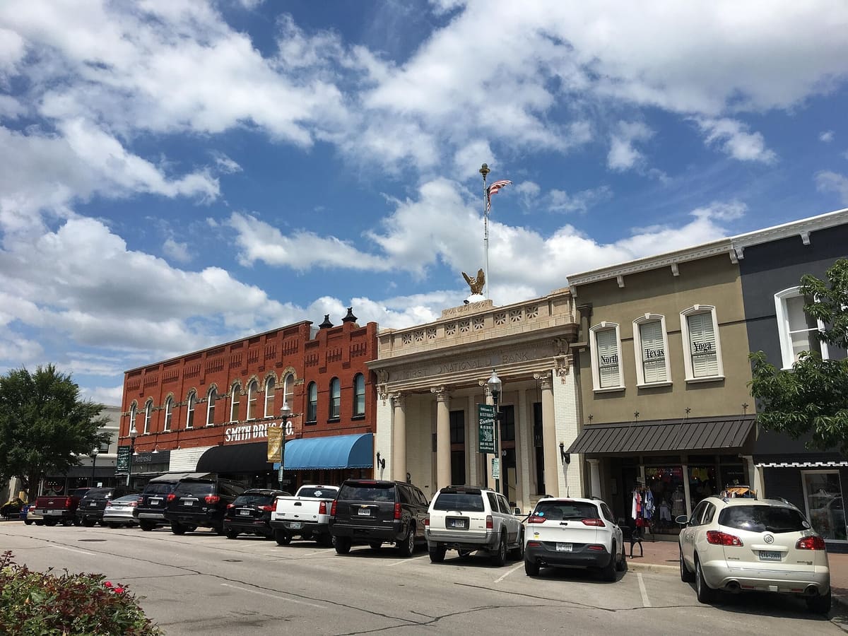 Storefronts on the McKinney square: a two-story red brick commercial block, the temple-front First National Bank building, and an Italianate two-part block