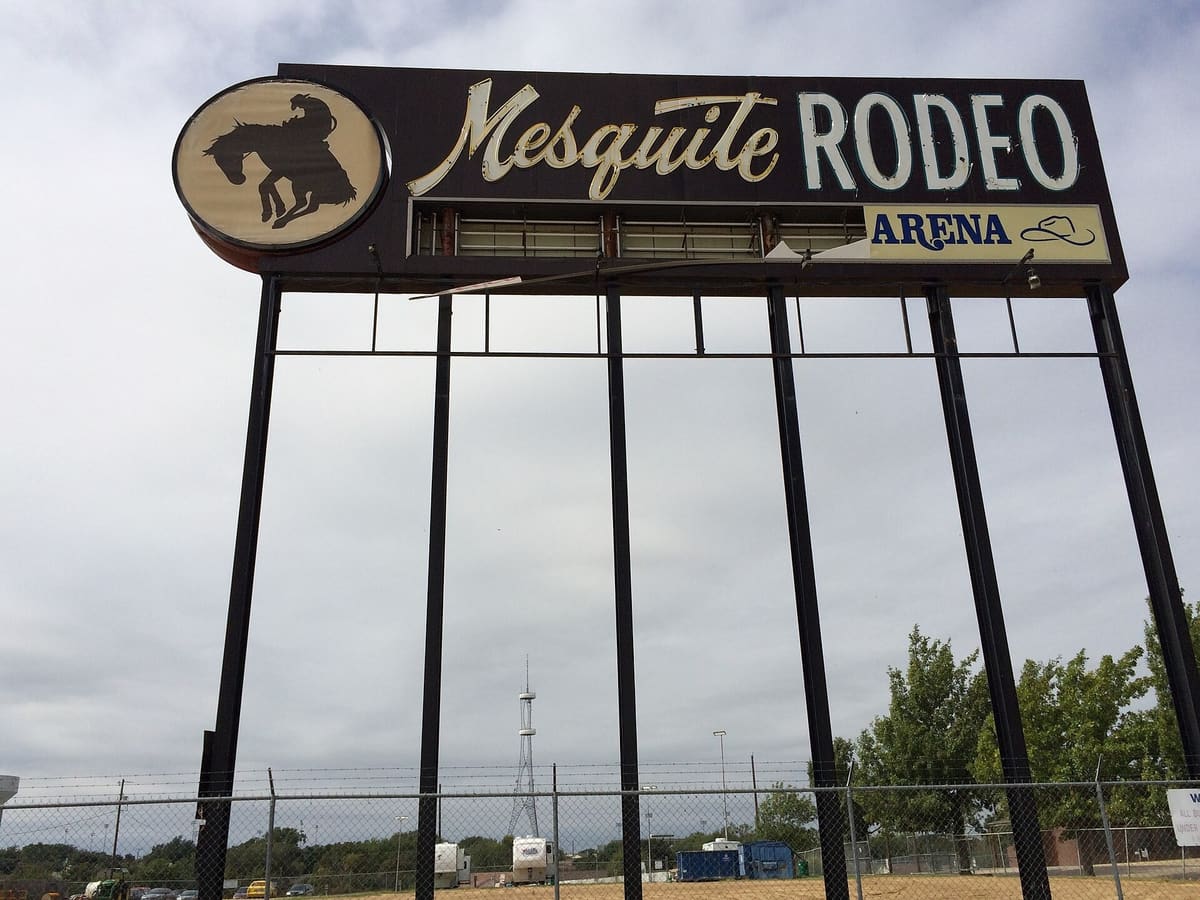 The Mesquite Rodeo Arena sign on tall black steel posts, with a bucking horse silhouette and neon script lettering against a gray sky