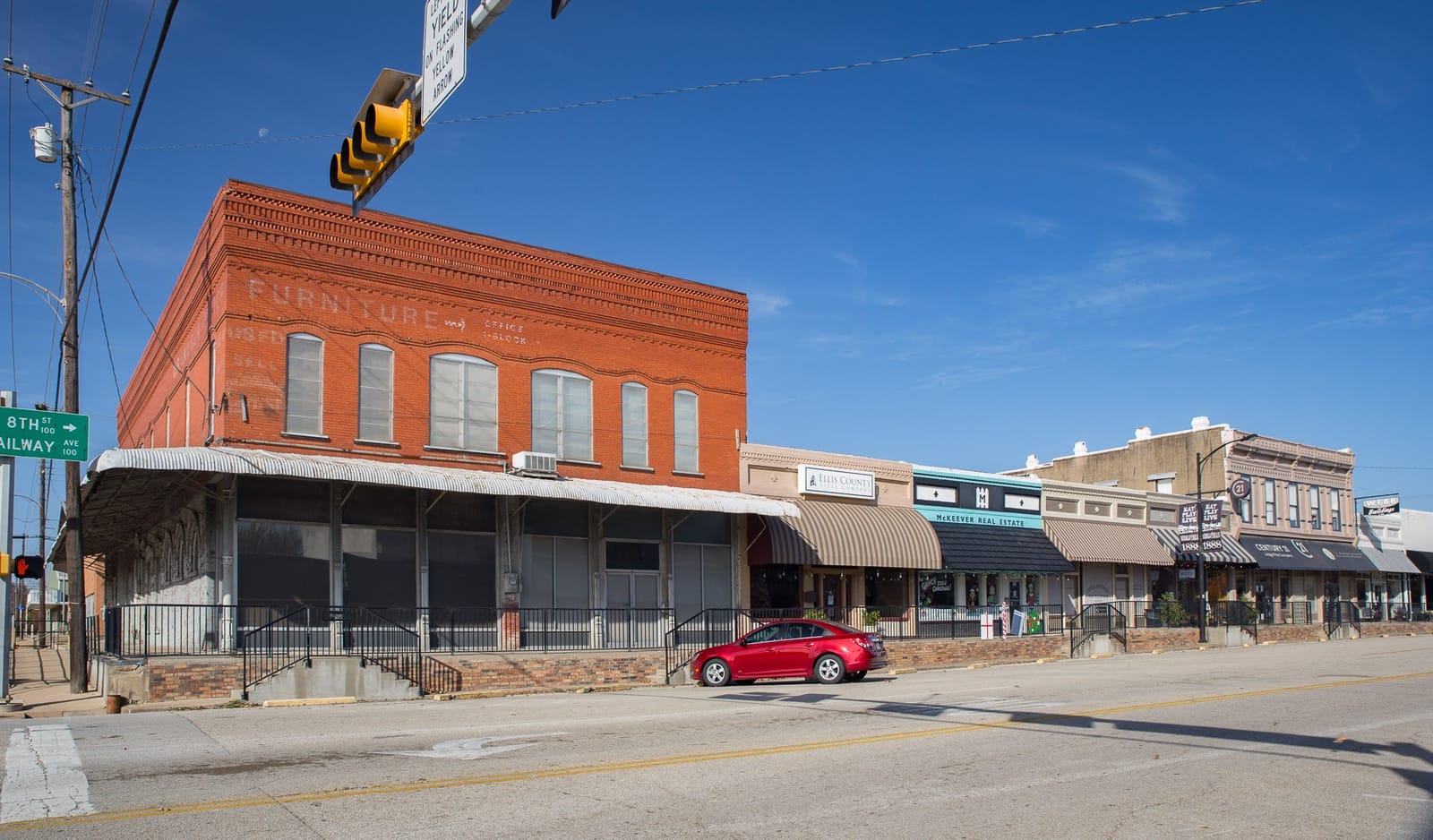 A two-story red brick building with a faded furniture sign and a row of awninged storefronts at 8th Street and Railway Avenue in downtown Midlothian, Texas