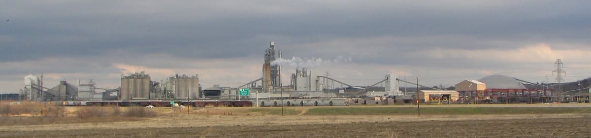 A long cement plant with silos, a preheater tower, conveyors, and a storage dome beside a highway in Midlothian, Texas under gray clouds