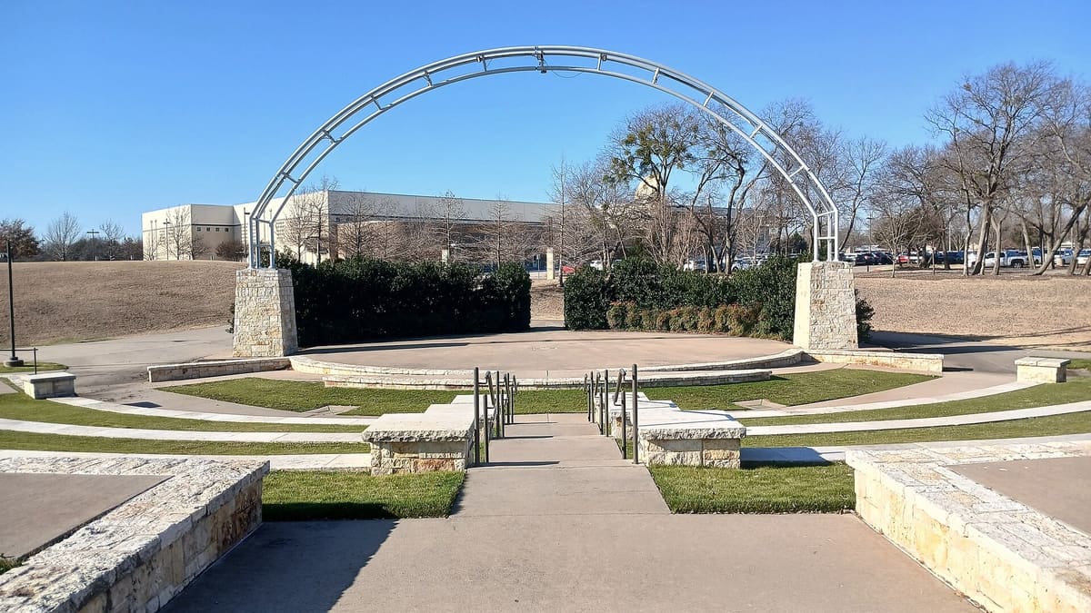 The amphitheater stage at Murphy Central Park in Murphy, Texas, under a steel arch on limestone piers, with stone seat walls and grass terraces in front