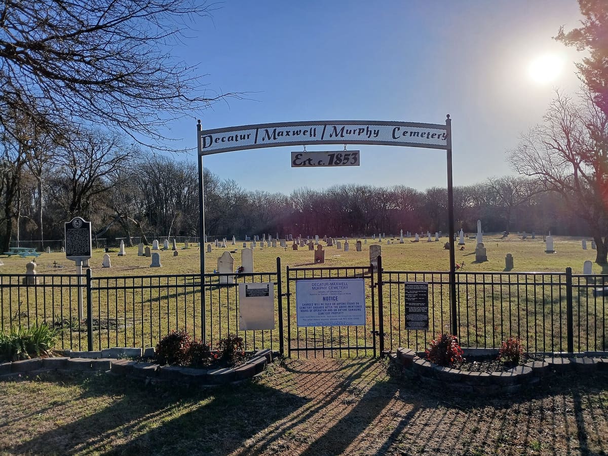 The arched iron entry sign of the Decatur Maxwell Murphy Cemetery in Murphy, Texas, with a historical marker and rows of old headstones behind a black fence