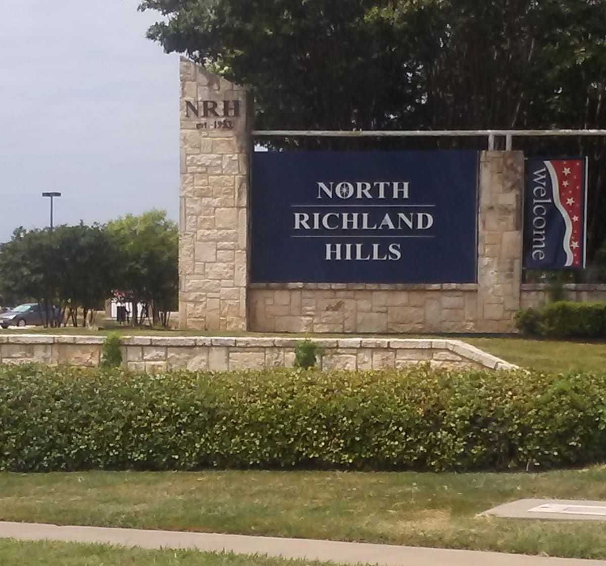 The North Richland Hills welcome monument on Boulevard 26, a limestone pillar carved NRH est. 1953 beside a navy sign panel and a red, white, and blue Welcome banner