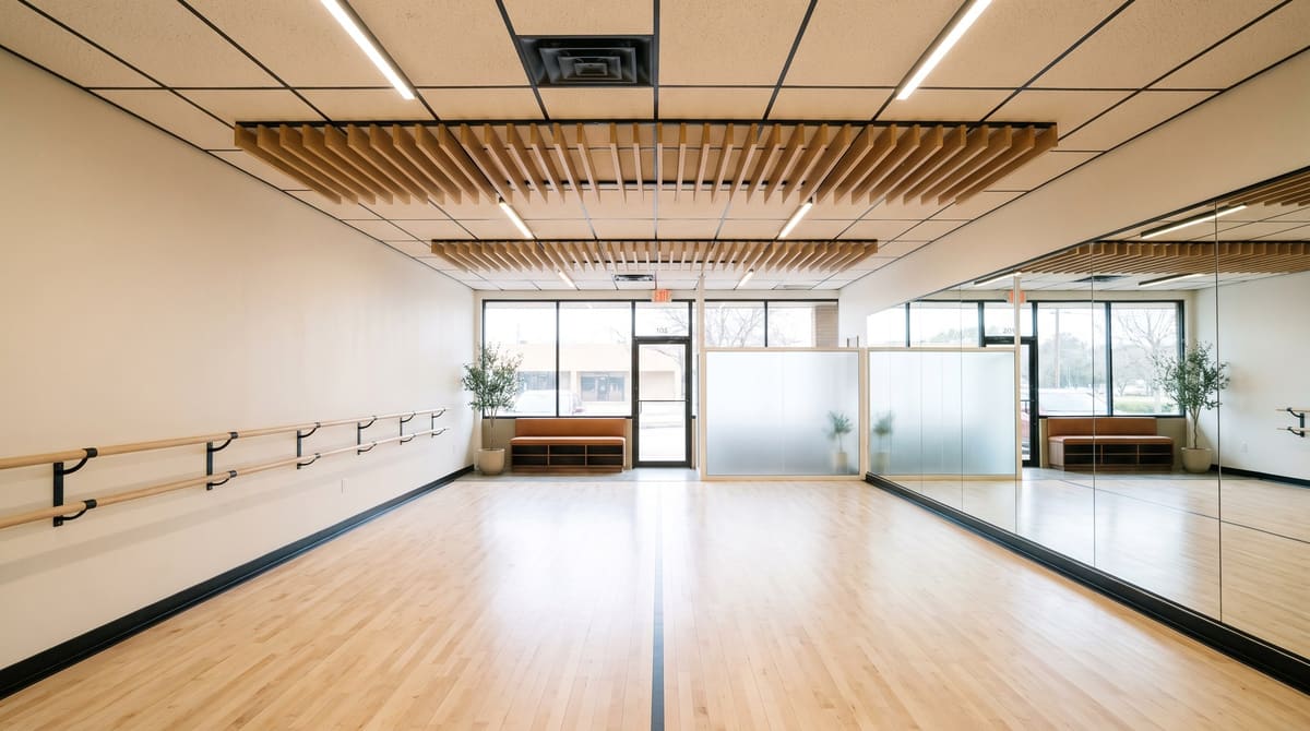 A finished dance studio in a retail suite with a pale maple sprung floor, a full mirror wall in thin black trim, ash ballet barres on black brackets, a white acoustic ceiling with wood baffles, and a small waiting bench by the storefront