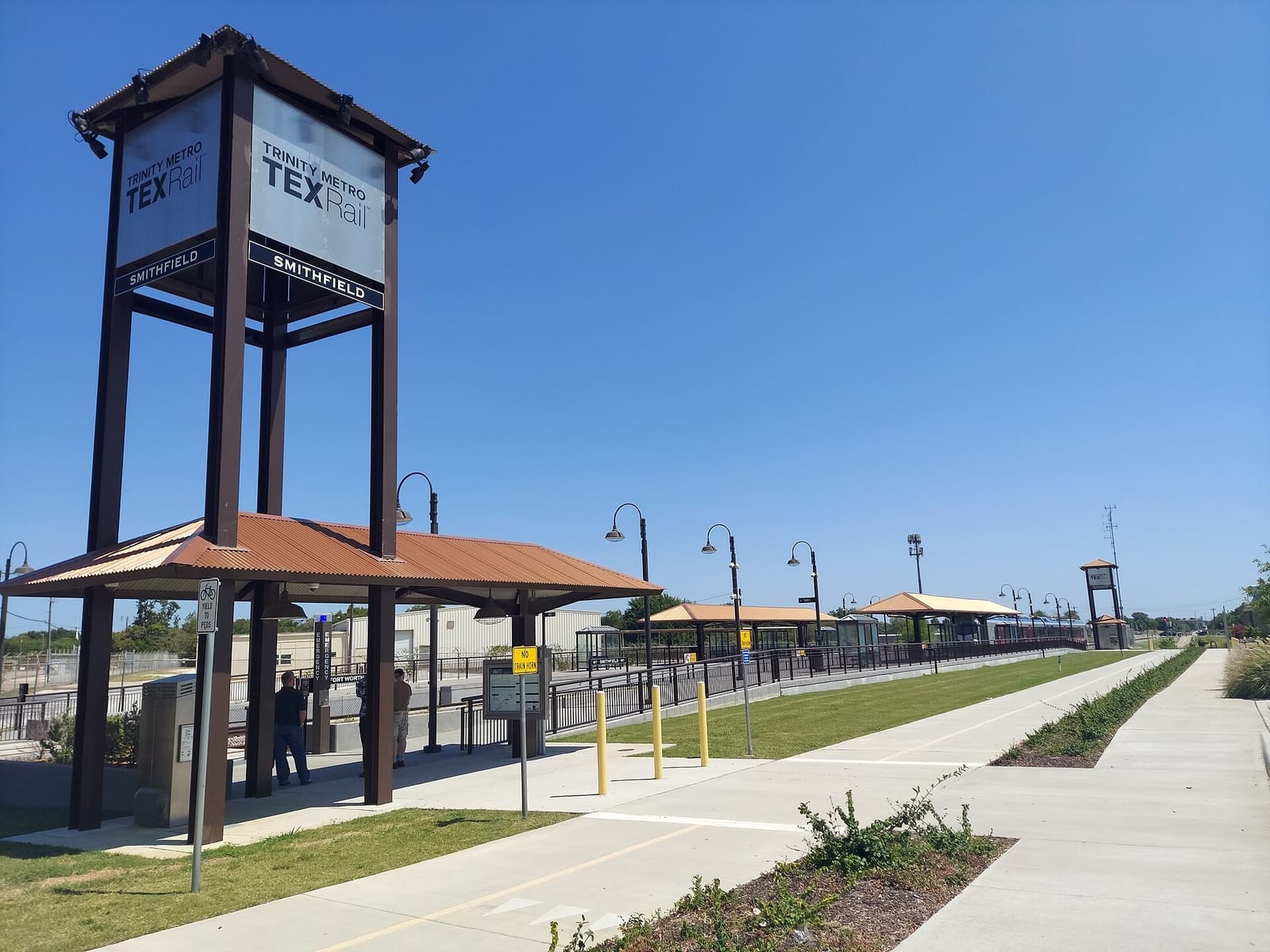 The TEXRail Smithfield station in North Richland Hills, a tall dark-framed sign tower reading Trinity Metro TEXRail Smithfield above a rust-colored metal shelter, with the platform stretching down the line under a clear blue sky
