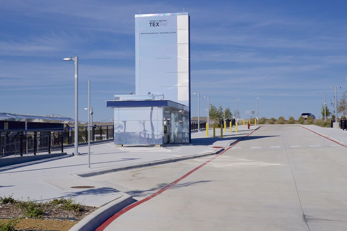 The North Richland Hills Iron Horse TEXRail station, a tall stainless steel sign tower labeled North Richland Hills Iron Horse Station above a small glass entry pavilion and a bus drive