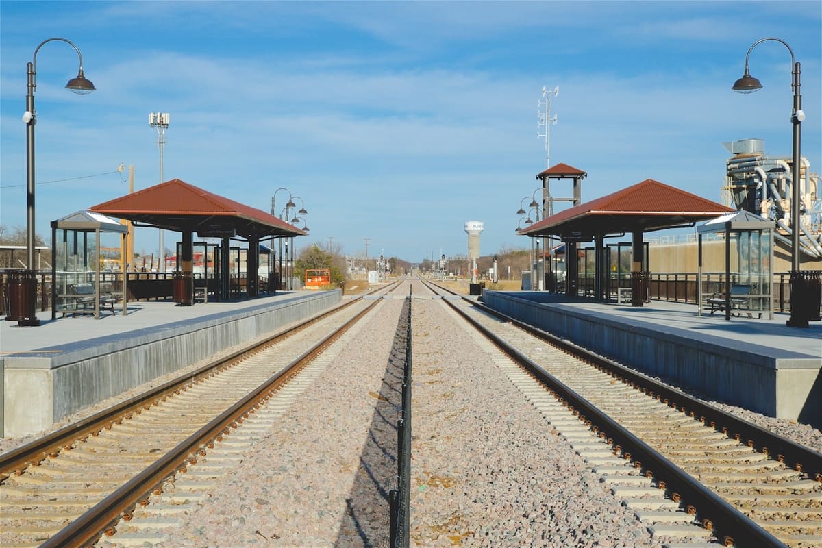 The TEXRail Smithfield station in North Richland Hills, two platforms with rust-red hip-roof shelters and curved lamp posts on either side of the tracks, with a white NRH water tower on the horizon