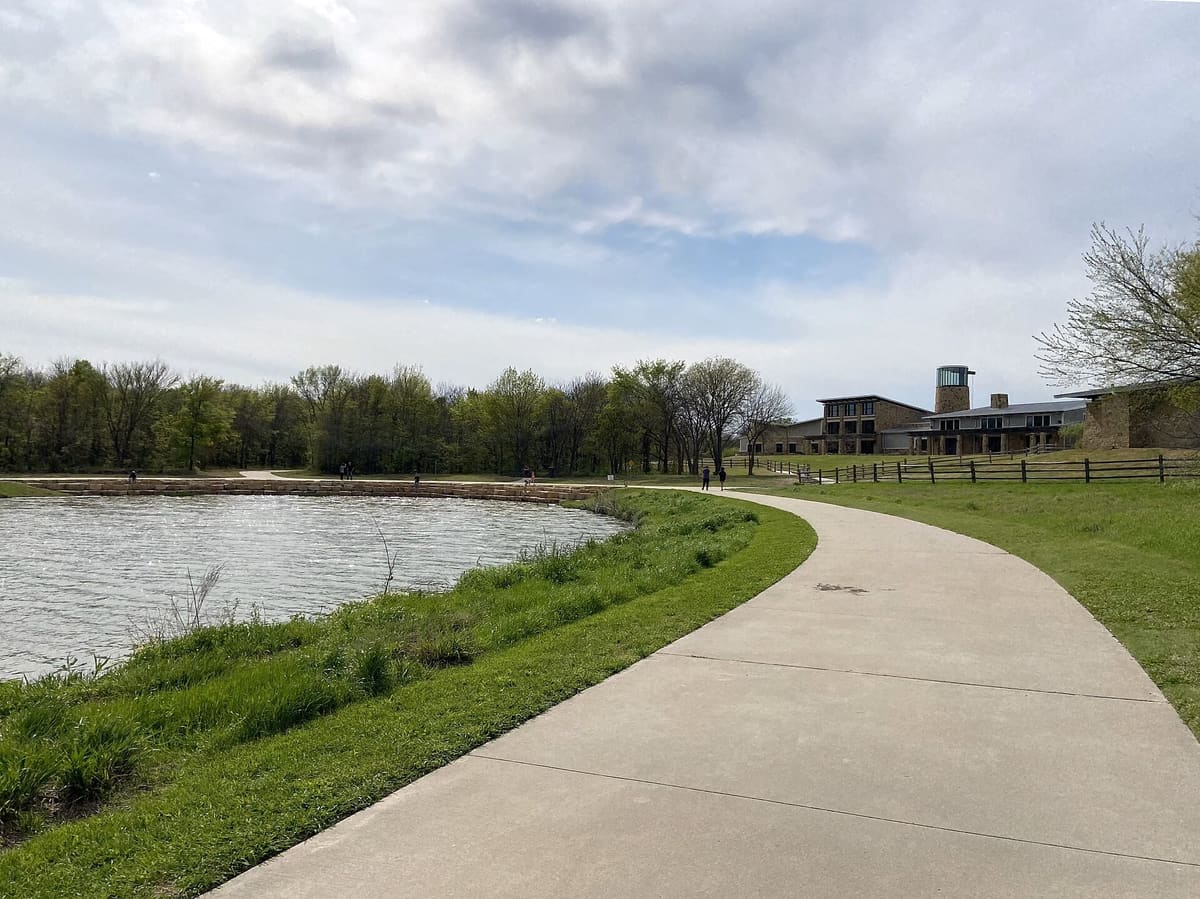 A concrete trail curving past a pond toward the stone Nature and Retreat Center at Oak Point Park in Plano