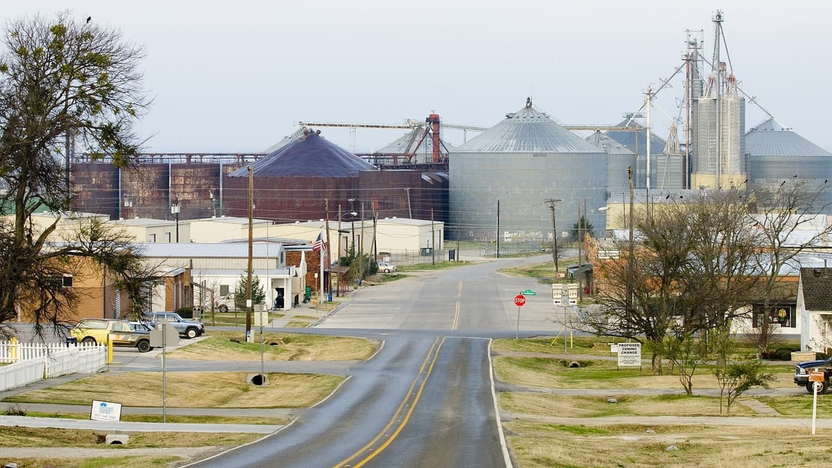 Downtown Prosper in 2008 with a two-lane road leading past low brick and metal buildings toward a cluster of grain silos