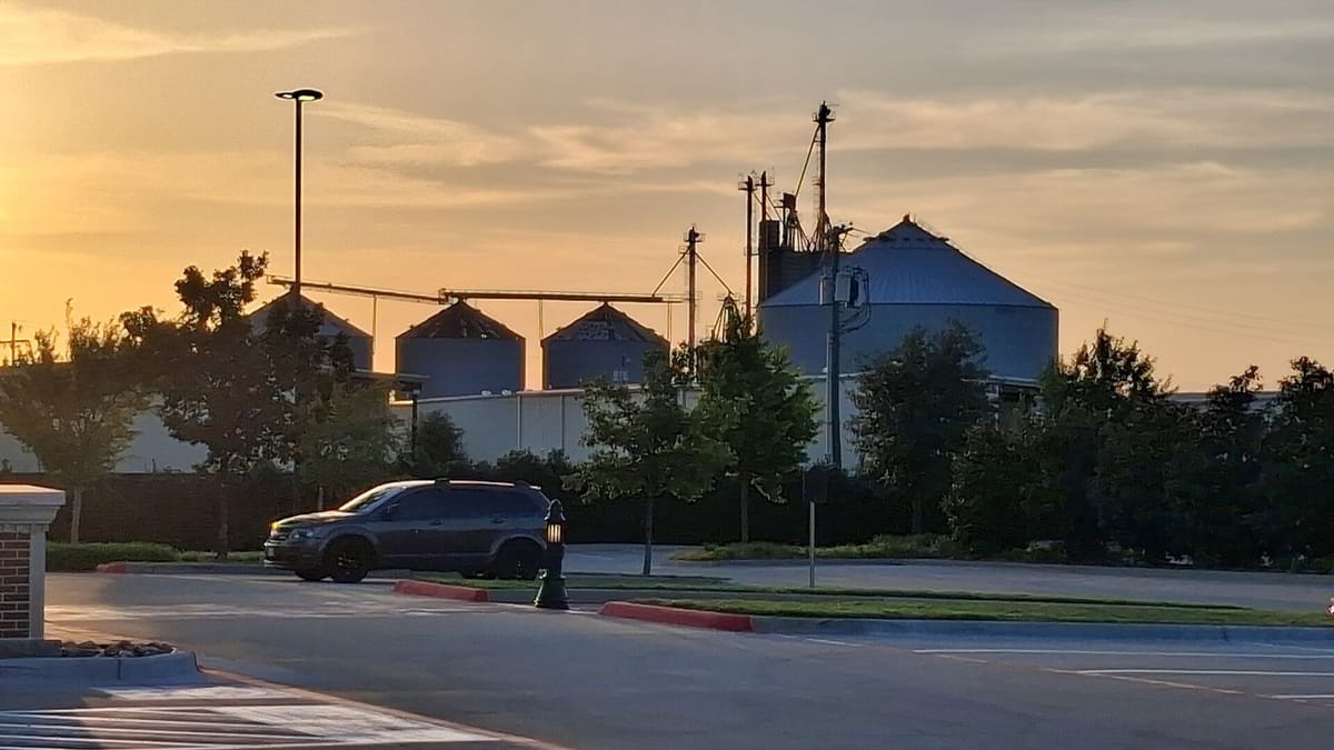 Downtown Prosper's grain silos and conveyor towers silhouetted against an orange sunset sky