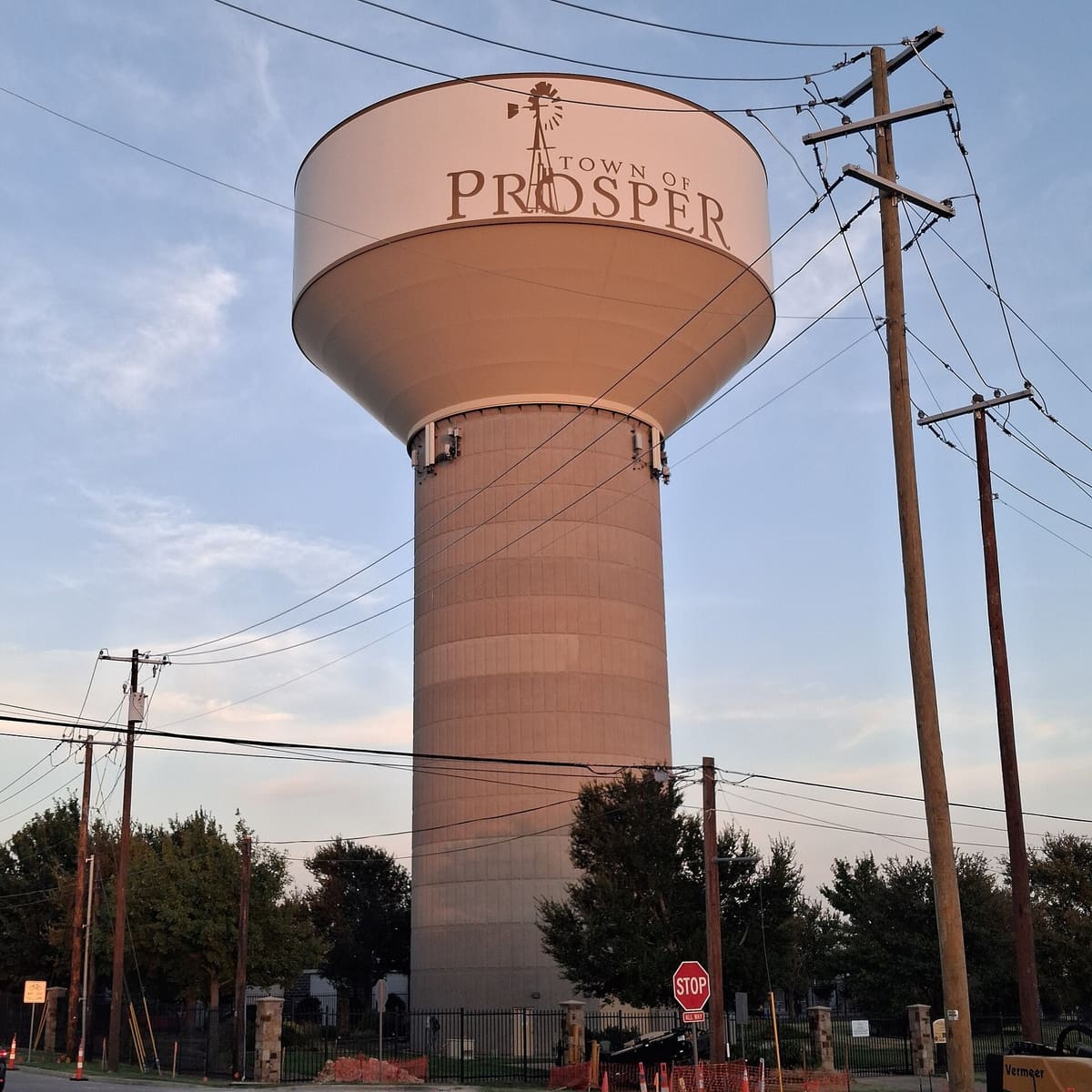 A tall water tower painted with the words Town of Prosper and a windmill, standing behind a fenced lot and power lines