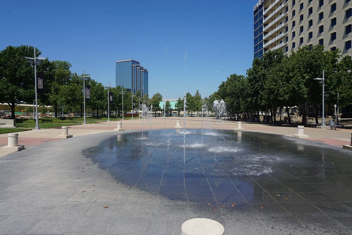 The Texas Instruments Foundation Community Fountain spraying water on the granite plaza at Galatyn Park in Richardson, with a hotel tower on the right and a glass office tower in the distance