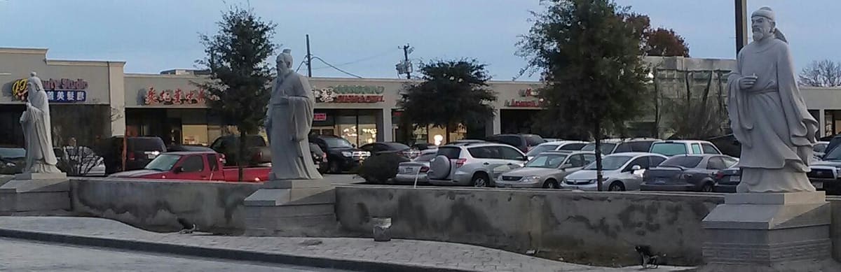 Stone statues of figures from Chinese history standing on pedestals in the square at DFW Chinatown in Richardson, with the shopping center storefronts and parking lot behind them