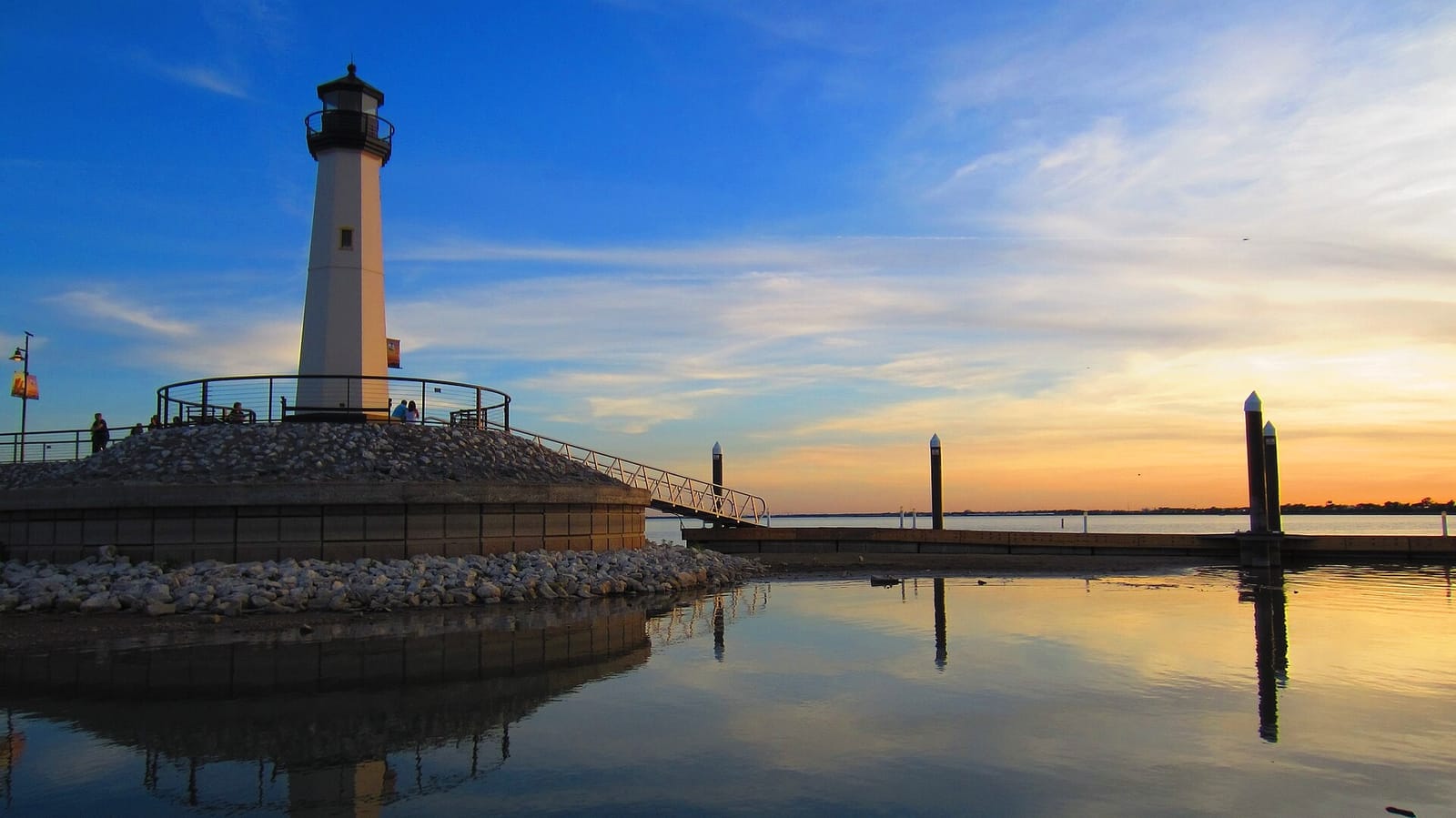 The white lighthouse on the waterfront at The Harbor in Rockwall, Texas, at sunset, with Lake Ray Hubbard calm and docks reflected in the water