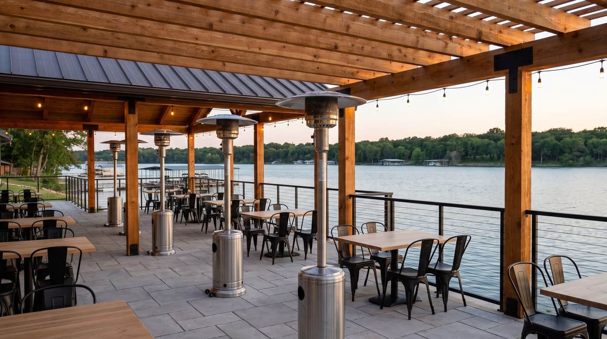 A covered lakeside restaurant terrace with a cedar pergola, tall stainless patio heaters, limestone pavers, and calm lake water beyond the railing