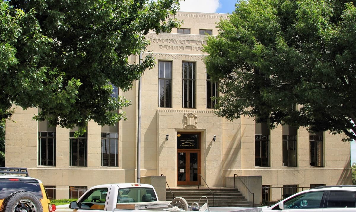 The limestone Art Deco Rockwall County Historic Courthouse at 101 East Rusk Street, with tall vertical windows and a carved panel over the entry, framed by oak trees