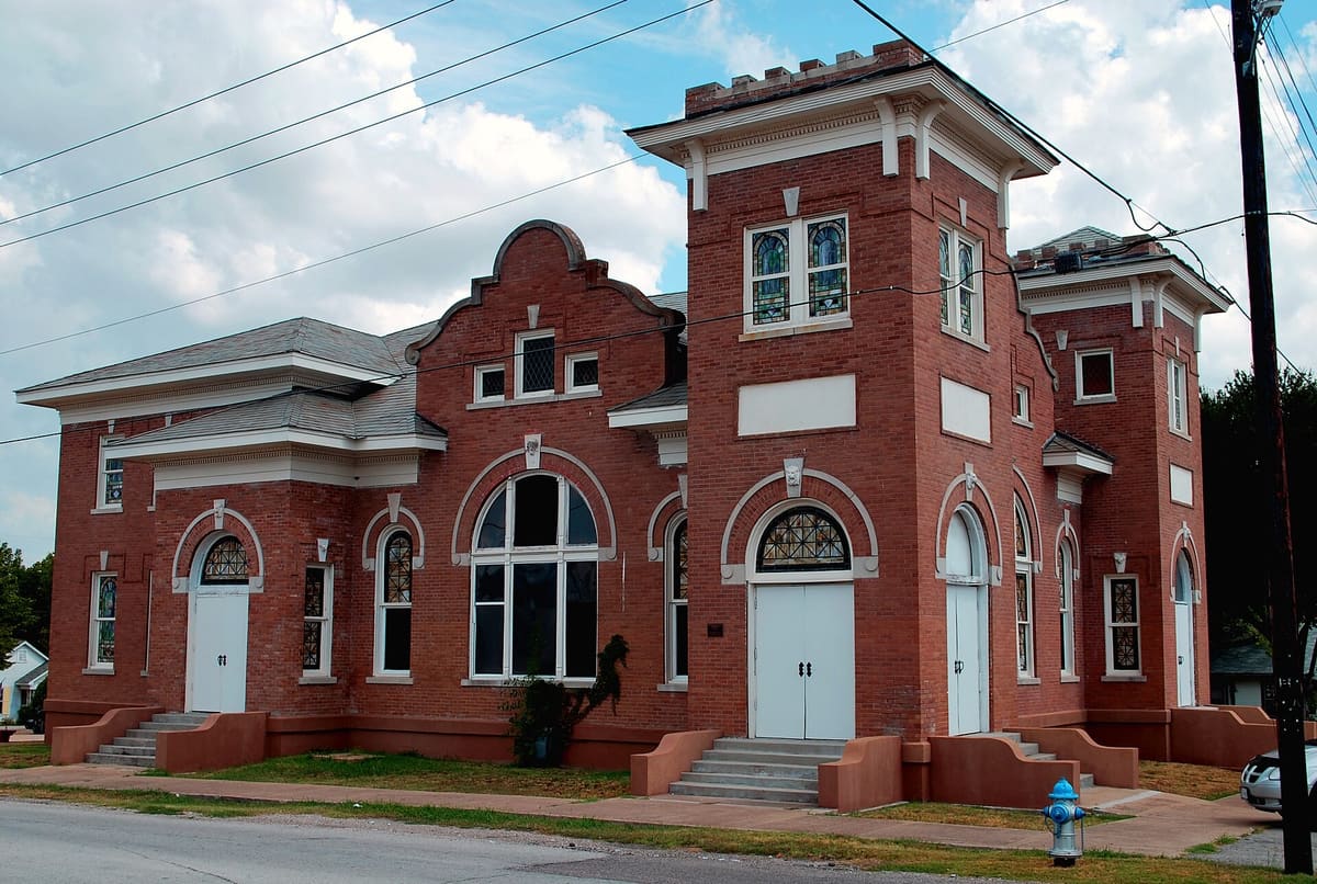 The red brick First Methodist Church of Rockwall with two towers, curved Mission Revival parapet, arched stained glass windows, and white double doors
