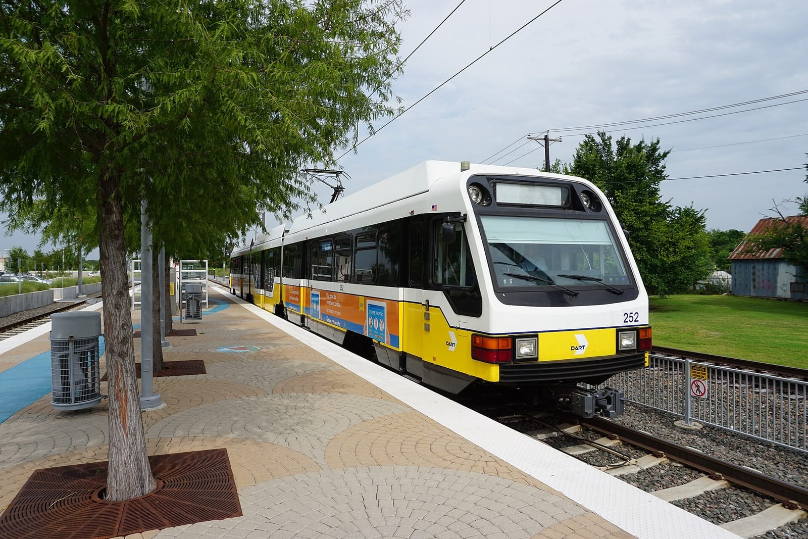 A yellow and white DART light rail train stopped at Downtown Rowlett Station beside a brick paver platform lined with young trees