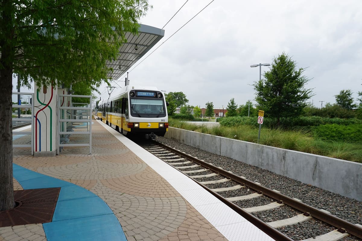 A DART Blue Line train arriving at the Downtown Rowlett Station platform under a curved metal canopy, with decorative rail-line art on a column