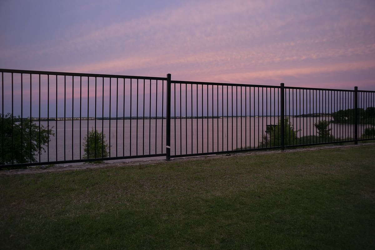 Lake Ray Hubbard at dusk under a pink and violet sky, seen through a black metal fence at the edge of a lawn