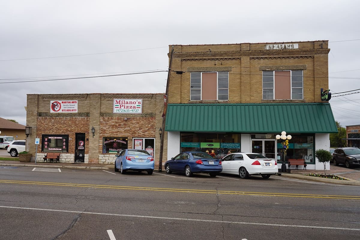 A two-story buff brick commercial building with a green standing seam awning and a faded W. F. Adams name on its parapet, beside one-story brick storefronts on Main Street in Royse City