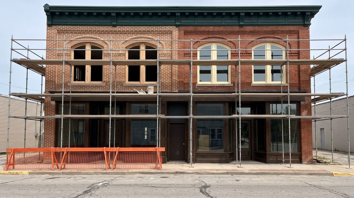 Scaffolding on an early 1900s two-story red brick downtown building with freshly repointed brick, restored wood upper windows, and a new dark green period storefront at street level