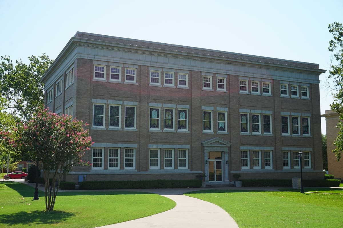 Sherman Hall at Austin College, a three-story tan brick building with a pedimented entrance, stained glass second floor windows, and a curving walk across a lawn