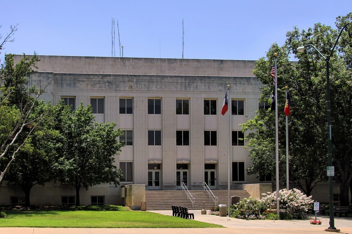 The pale limestone Grayson County Courthouse in Sherman with fluted pilasters, Grayson County Courthouse lettering, front steps, and flagpoles framed by trees