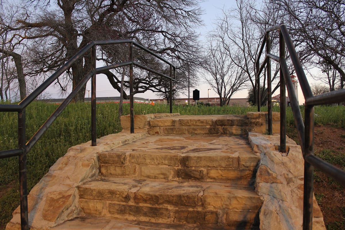Rough sandstone steps with black iron handrails climbing a grassy rise to the Thomas Easter Cemetery in Southlake, Texas, with bare trees and a shopping center behind