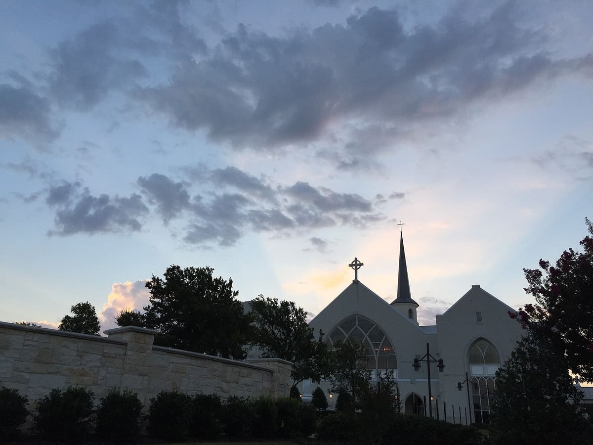 White's Chapel United Methodist Church in Southlake at dusk, with a pale gabled sanctuary, a tall steeple, a Celtic cross, and a cut stone wall in the foreground