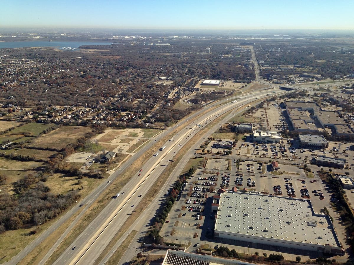 Aerial view of State Highway 114 crossing Southlake, Texas, with frontage roads, big-box stores and parking lots on one side and wooded neighborhoods on the other
