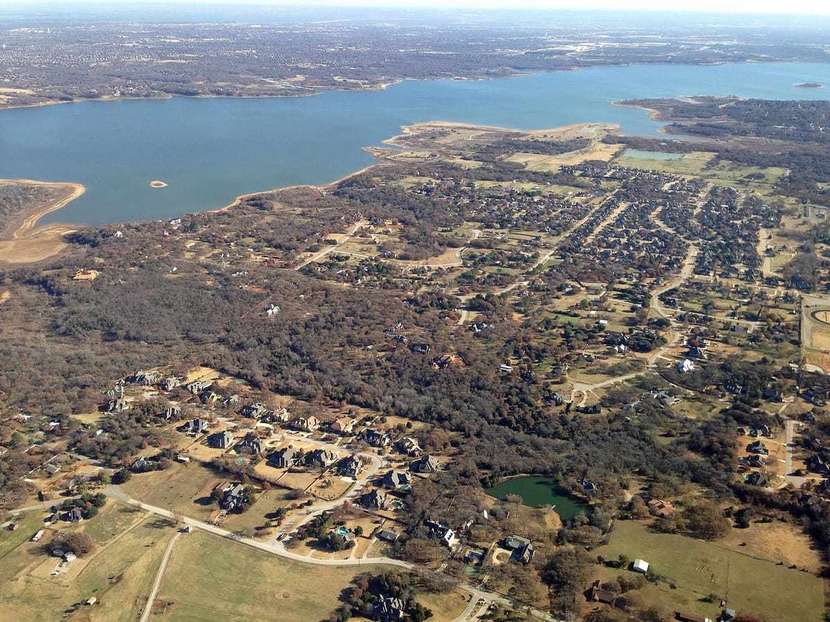 Aerial view of part of Southlake, Texas, with large wooded lots, a curving subdivision street of big homes, a pond, and Grapevine Lake on the horizon